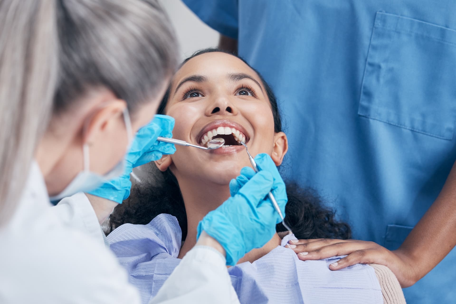 A woman receives a dental check-up, smiling with her mouth open. A dentist in gloves holds dental tools, assisted by a person in blue scrubs who gently places a hand on her shoulder, creating a supportive environment.